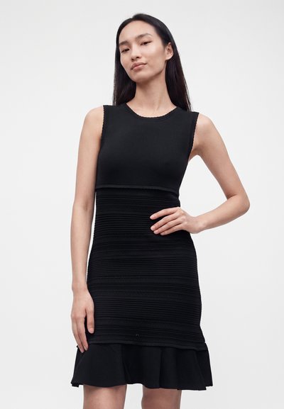 Femme aux longs cheveux foncés portant une robe noire sans manches avec une partie centrale texturée et un ourlet à volants, posant avec une main sur la taille.