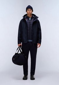 Marineblauwe waterdichte jas over een marineblauwe gewatteerde bodywarmer, gecombineerd met een donkere broek en zwarte schoenen. Houdt een donkere weekendtas vast en draagt een muts.