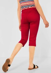 Pantalones capri rojos con un diseño ajustado, cintura de altura media y dobladillo con puños. El material parece suave y cuenta con dos bolsillos traseros. Llevado con sandalias negras.