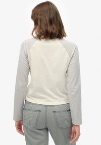 Vrouw met schouderlang bruin haar draagt een crème- en grijskleurig shirt met lange mouwen en gestreepte broek, gezien van achteren tegen een witte achtergrond.