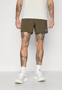 Athletic shorts in olive green, made of lightweight fabric. Features a white logo on the hem. Model wears white athletic shoes and crew socks.