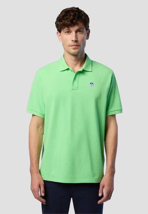 Uomo con capelli ricci che indossa una polo a maniche corte verde chiaro con un piccolo logo rotondo sul lato sinistro del petto, in piedi davanti a uno sfondo grigio.