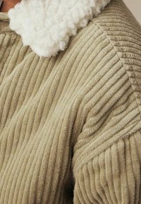 Vêtement en velours côtelé vert olive texturé avec un col blanc duveteux, présentant des motifs à nervures verticales et un design à coupe décontractée.