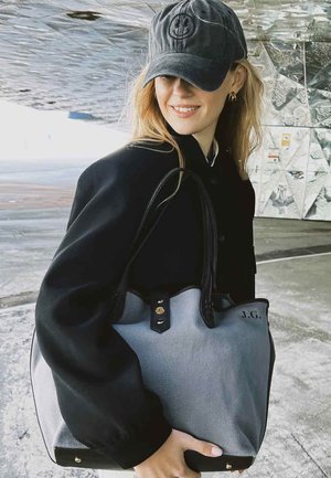 Sac fourre-tout gris et noir avec une texture lisse, un haut arrondi, des accents en cuir noir et des éléments en métal doré. Portant les initiales "J.G." sur le côté.