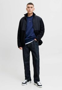 Black fleece jacket with high collar over a navy sweatshirt, paired with dark denim jeans and black and white sneakers.