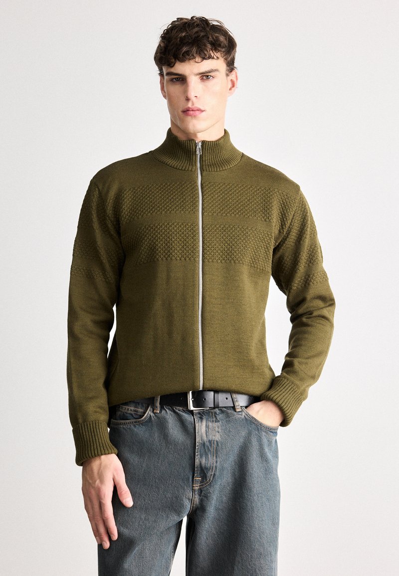 Olive green knitted zip-up sweater with ribbed collar and cuffs, featuring textured pattern stripes across the chest and a smooth finish.