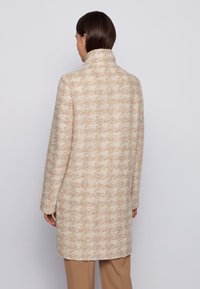 Kvinde med mørkt hår iført en beige og cremefarvet lang frakke med hundetandsmønstret tekstur, set bagfra mod en ensartet lys baggrund.