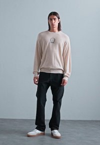 Beige stickad tröja med rund halsringning och logotypdetalj, i kombination med svarta byxor och vita sneakers. Mjuk textur och avslappnad passform.