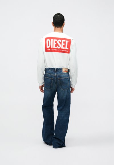 Мъж в бяла риза с дълги ръкави и червен логотип "DIESEL" на гърба, облечен в сини дънки, застава с гръб към бял фон.