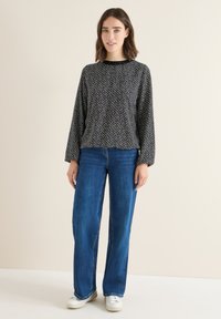 Maglia a maniche lunghe con motivo in bianco e nero abbinata a jeans blu a gamba larga e sneakers bianche, in piedi contro uno sfondo neutro.