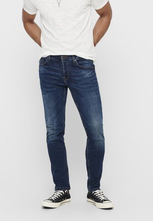 Mand iført mørkeblå slim fit jeans, sort-hvide sneakers og en hvid kortærmet t-shirt, står med hænderne bag ryggen.