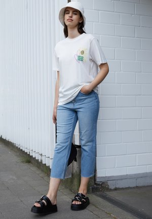 Camiseta branca com design de espiral multicolorido e texto, calças jeans cropped de tom azul claro, e sandálias pretas chunky com tiras. Chapéu de balde branco.
