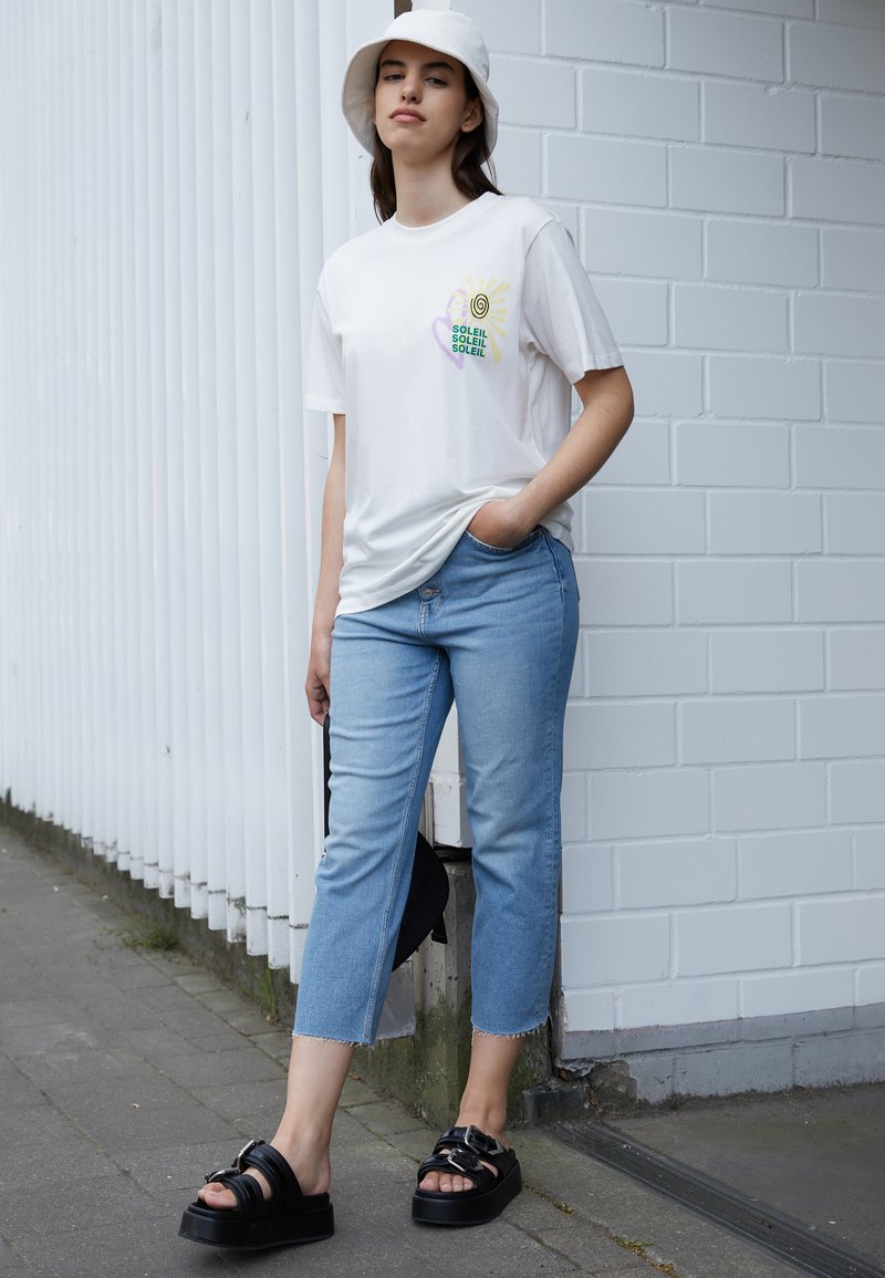Camiseta branca com design de espiral multicolorido e texto, calças jeans cropped de tom azul claro, e sandálias pretas chunky com tiras. Chapéu de balde branco.