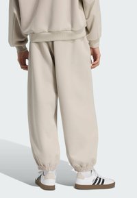 Béžové voľné tepláky s elastickými manžetami, v kombinácii s bielymi teniskami s čiernymi pruhmi. Hladká textura, voľný strih a ležérny dizajn.