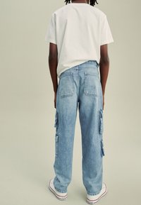 Világoskék farmer cargo nadrág oldal zsebekkel, laza szabással és varrott részletekkel. Egyszerű fehér pólóval és fehér tornacipővel párosítva.