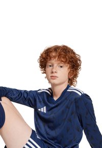 Maglia atletica blu navy con motivi geometrici neri, con strisce bianche sulle spalle e colletto a V. Bambino seduto con espressione neutra.