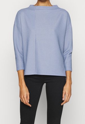 Frau trägt ein blau-weiß gestreiftes Langarmoberteil mit vertikalen und horizontalen Mustern, kombiniert mit schwarzer Hose.