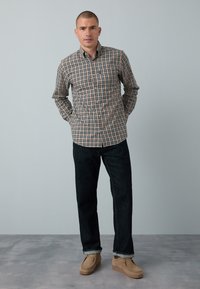 Chemise à carreaux beige, bleue et marron, associée à un jean en denim foncé et des chaussures marron clair. La coupe est décontractée et moderne.