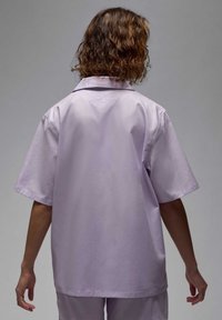 Camicia a maniche corte lavanda con vestibilità rilassata, dotata di colletto e un piccolo logo ricamato sulla parte superiore della schiena, realizzata in tessuto morbido.