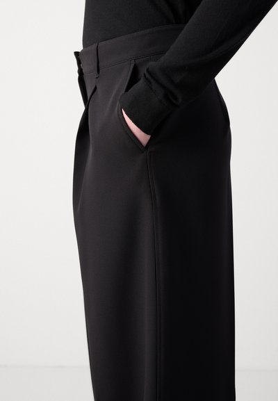 Pantalons noirs avec un tissu lisse et texturé, dotés de poches latérales et d'une coupe ajustée, présentés de côté sur un fond neutre.