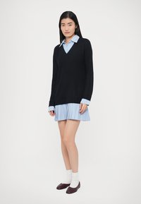 Svart strikket V-hals genser lagd over en lyseblå skjorte med krage og vertikale striper. Modellen har på seg et plisset skjørt og mørke flate sko.