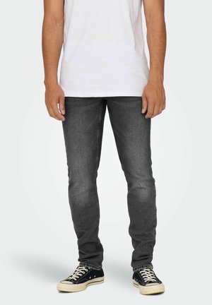 Mężczyzna w białej koszulce, ciemnoszarych jeansach typu slim fit oraz czarnych niskich trampkach stoi na jednolitym tle.