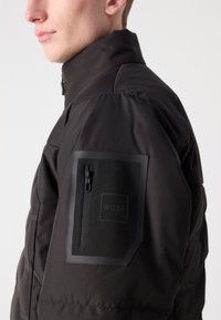 Crna puf jakna s visokim ovratnikom, koja ima zippered džep na lijevoj ruci. Teksturalni detalji s "BOSS" brendingom.