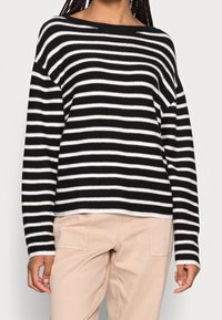 Černobílý pruhovaný svetr s širokým výstřihem, dlouhými rukávy a volným střihem. Látka působí měkce a má pletenou texturu.