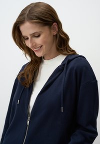 Felpa con zip di un blu navy, realizzata in morbido tessuto a maglia a coste, con cappuccio regolabile e zip argentata. Indossata sopra una maglietta bianca a girocollo.