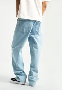 Jeans de mezclilla en azul claro con corte holgado, diseño de cinco bolsillos y textura descolorida. La persona que los lleva puestos está con zapatillas negras, orientándose ligeramente hacia un lado.