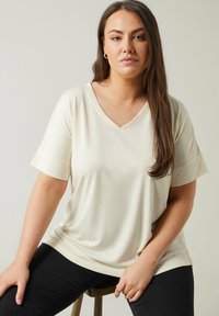 Khaki V-paita, joka on valmistettu sileästä kankaasta, lyhyet hihat ja rento leikkaus. Yhdistetty tummiin farkkuihin, istuu puuistuimella.