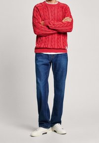 Roter Kabelstrickpullover mit Rundhalsausschnitt, kombiniert mit blauen Jeans und weißen Sneakern. Der Pullover hat gerippte Bündchen und einen gerippten Saum.