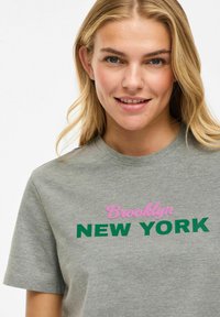 T-shirt en coton gris présentant le texte "Brooklyn" en rose et "NEW YORK" en vert. Il a des manches courtes et un col rond.