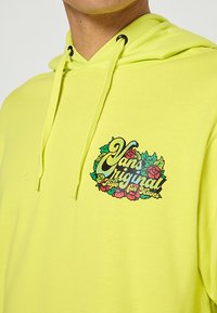 Sweat à capuche jaune vif en coton, présentant un motif à l'avant avec le texte "Vans Original" entouré de motifs floraux. Capuche avec cordon de serrage ajustable.