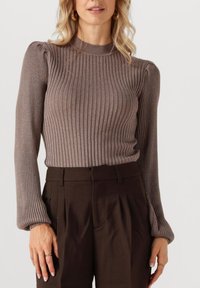 Brauner Rippstrickpullover mit langen Ärmeln, der ein tailliertes Design und puffy Schulterdetails aufweist. Kombiniert mit hochgeschnittenen, dunkelbraunen Hosen.