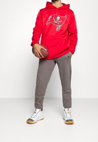 Haut à capuche rouge avec le logo des Buccaneers, pantalon de survêtement gris avec une bande latérale, et baskets blanches avec des accents jaunes. Tenant un ballon de football.