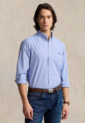 Νέος άνδρας με μεσαίου μήκους σκούρα μαλλιά, ντυμένος με μπλε καρό πουκάμισο με κουμπιά και ριγμένα μανίκια, σκούρο τζιν, καφέ ζώνη και ρολόι στον καρπό.