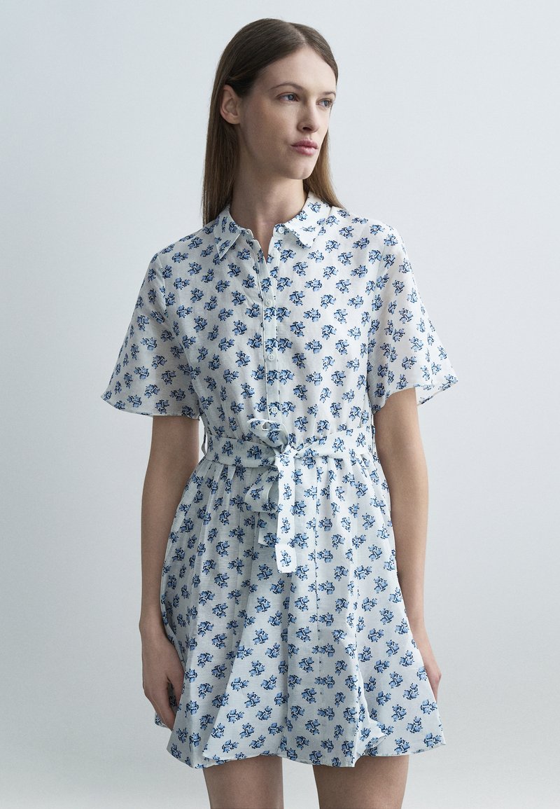 Γυναίκα που φοράει λευκό φόρεμα με κοντά μανίκια, διακοσμημένο με μπλε λουλουδάτα σχέδια και ζώνη στη μέση, στέκεται μπροστά σε απλό ανοιχτό φόντο.