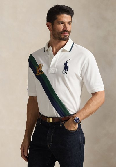 Camiseta tipo polo blanca con una franja diagonal verde y azul marino, logo bordado, mangas cortas, combinada con jeans oscuros y un cinturón marrón.