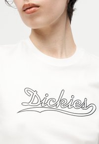 Κοντινό πλάνο ενός ατόμου που φοράει λευκό πουκάμισο με το λογότυπο "Dickies" σε μαύρο χρώμα στο στήθος, με τα χείλη ελαφρώς ανοιχτά.