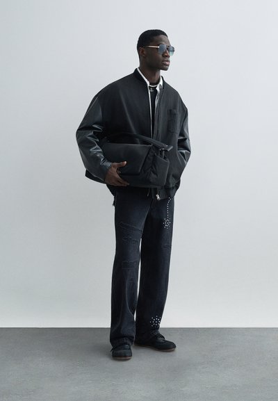 Férfi fekete dzsekiben, kopott fekete farmernadrág gyöngyös részletekkel, napszemüvegben, fekete táskát tartva, egyszerű szürke háttér előtt áll.