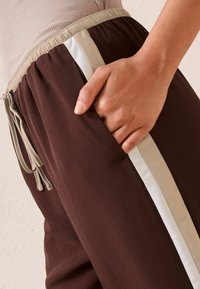 Bruna joggers i ett mjukt tyg har en beige dragsko och sidostrick. Midjebandet är elastiskt och det finns en hand i en sidoficka.