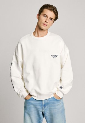 Sweatshirt blanc surdimensionné avec un col ras du cou côtelé, arborant un logo bleu foncé sur la poitrine et la manche. Associé à un jean bleu clair.