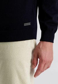 Marine gebreide trui met geribde manchetten, voorzien van een klein metalen label nabij de zoom. Een hand die zich nabij beige corduroy broek bevindt.
