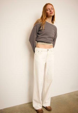 Jonge vrouw met een zwart-wit gestreept crop top met lange mouwen, witte wijde broekspijpen en bruine platte schoenen, staand tegen een witte muur.