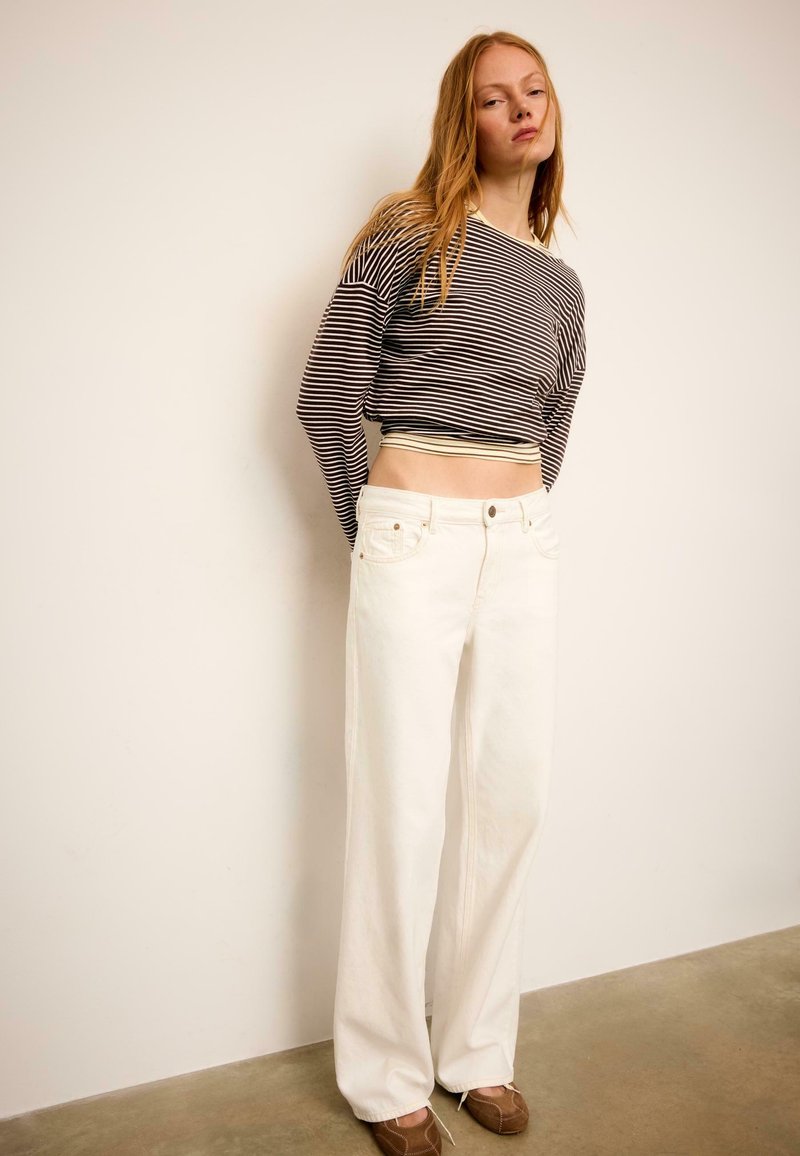 Jeune femme portant un crop top à manches longues rayé noir et blanc, un pantalon blanc à jambes larges, et des chaussures plates marron, debout contre un mur blanc.