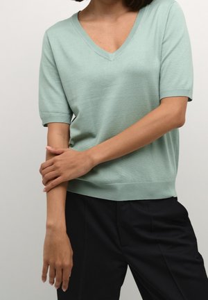 Kobieta ubrana w jasnozielony sweter z krótkim rękawem i dekoltem w serek oraz czarne spodnie, z lewą ręką dotykającą prawego przedramienia.