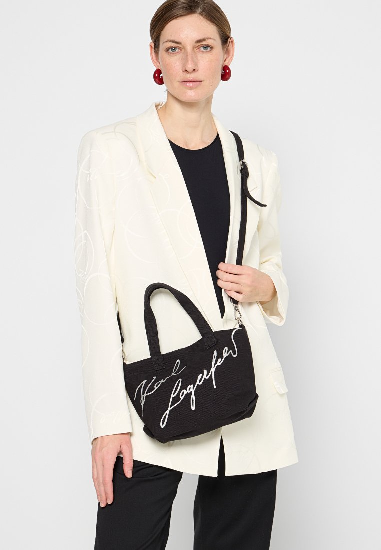 Sac à main en tissu noir avec deux poignées courtes, logo signature blanc, forme rectangulaire et bandoulière amovible.