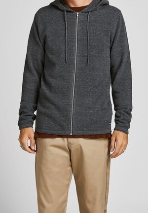 Personne portant un sweat à capuche zippé gris foncé avec cordons par-dessus une chemise marron et un pantalon beige, les bras détendus le long du corps.