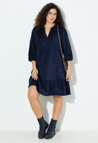 Nainen, jolla on kihara hiukset, on pukeutunut tummansiniseen polvipituiseen mekkoon, mustiin nauhakenkiin ja hän kantaa oliivinvihreää olkalaukkua seisten valkoista seinää vasten.